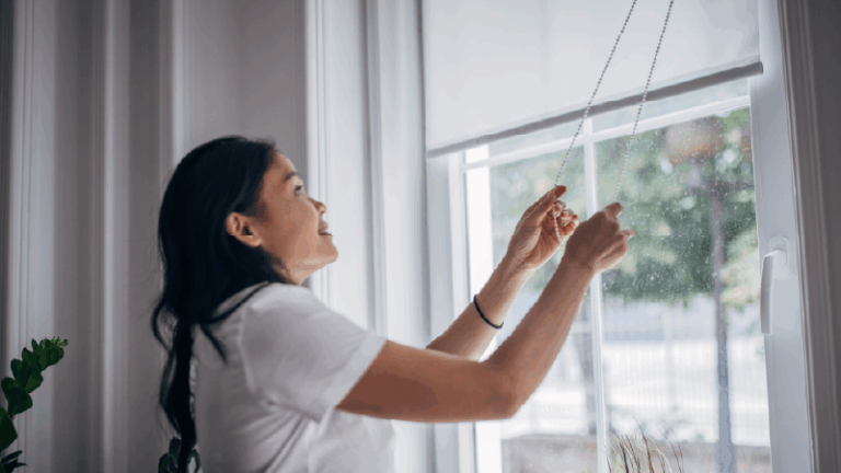 a woman standing by a window adjusting the blinds